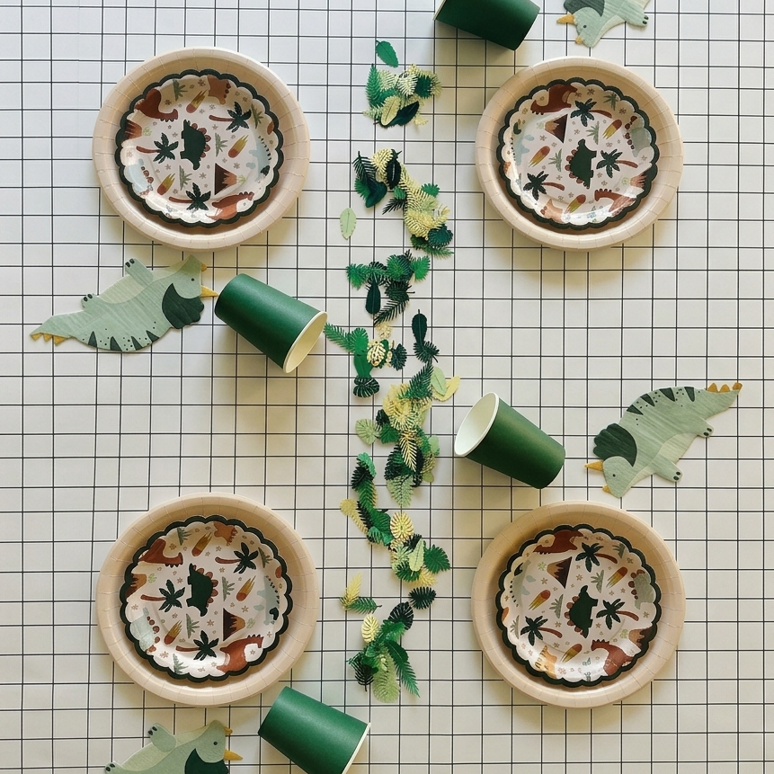 Vista aérea de una mesa decorada para fiesta infantil con platos, vasos, servilletas de dinosaurios y confeti de hojas sobre un mantel de papel cuadriculado.