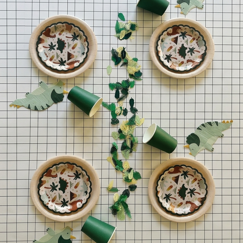 Vista aérea de una mesa decorada para fiesta infantil con platos, vasos, servilletas de dinosaurios y confeti de hojas sobre un mantel de papel cuadriculado.