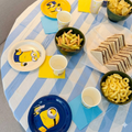 Decoración de mesa para fiesta infantil de los Minions con platos de papel, vasos, servilletas y boles de aperitivos con patatas fritas y sándwiches sobre un mantel a rayas.