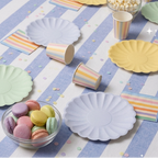 Primer plano angular de la mesa de cumpleaños con platos de papel verde menta y azul lavanda, vasos de rayas de colores pastel de pie, un cuenco con palomitas y otro con macarons, decorado con confeti delicado.