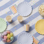 Primer plano angular de la mesa de cumpleaños con platos de papel verde menta y azul lavanda, vasos de rayas de colores pastel de pie, un cuenco con palomitas y otro con macarons, decorado con confeti delicado.