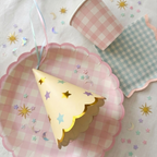 Primer plano de menaje para fiesta infantil de hadas con plato rosa de cuadros vichy, vaso verde pastel y gorrito de fiesta color crema con estrellas doradas y bordes festoneados sobre mantel blanco.