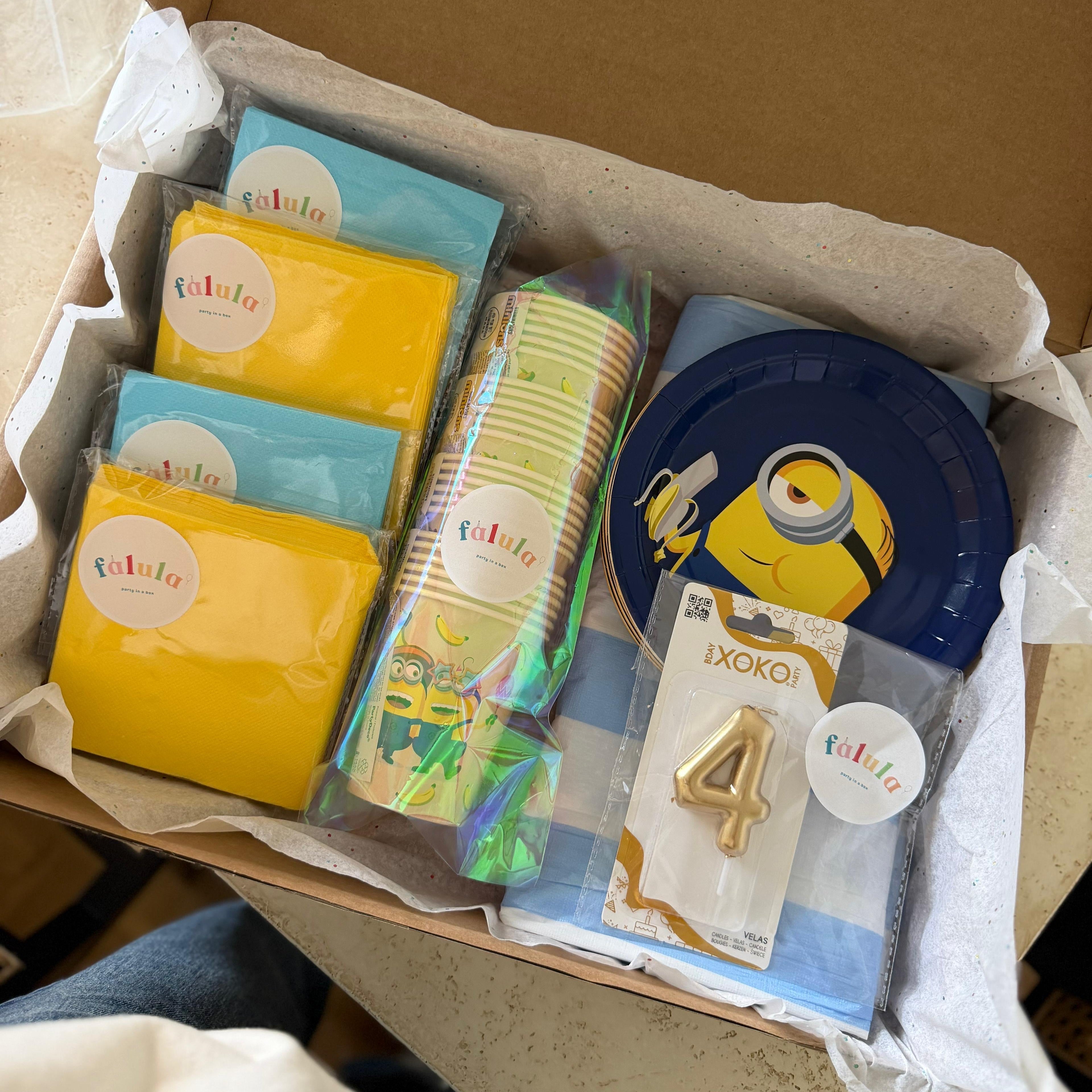 Caja de cartón abierta que contiene un set completo de fiesta de los Minions con platos, vasos, servilletas amarillas y azules, y una vela de cumpleaños con el número 4.