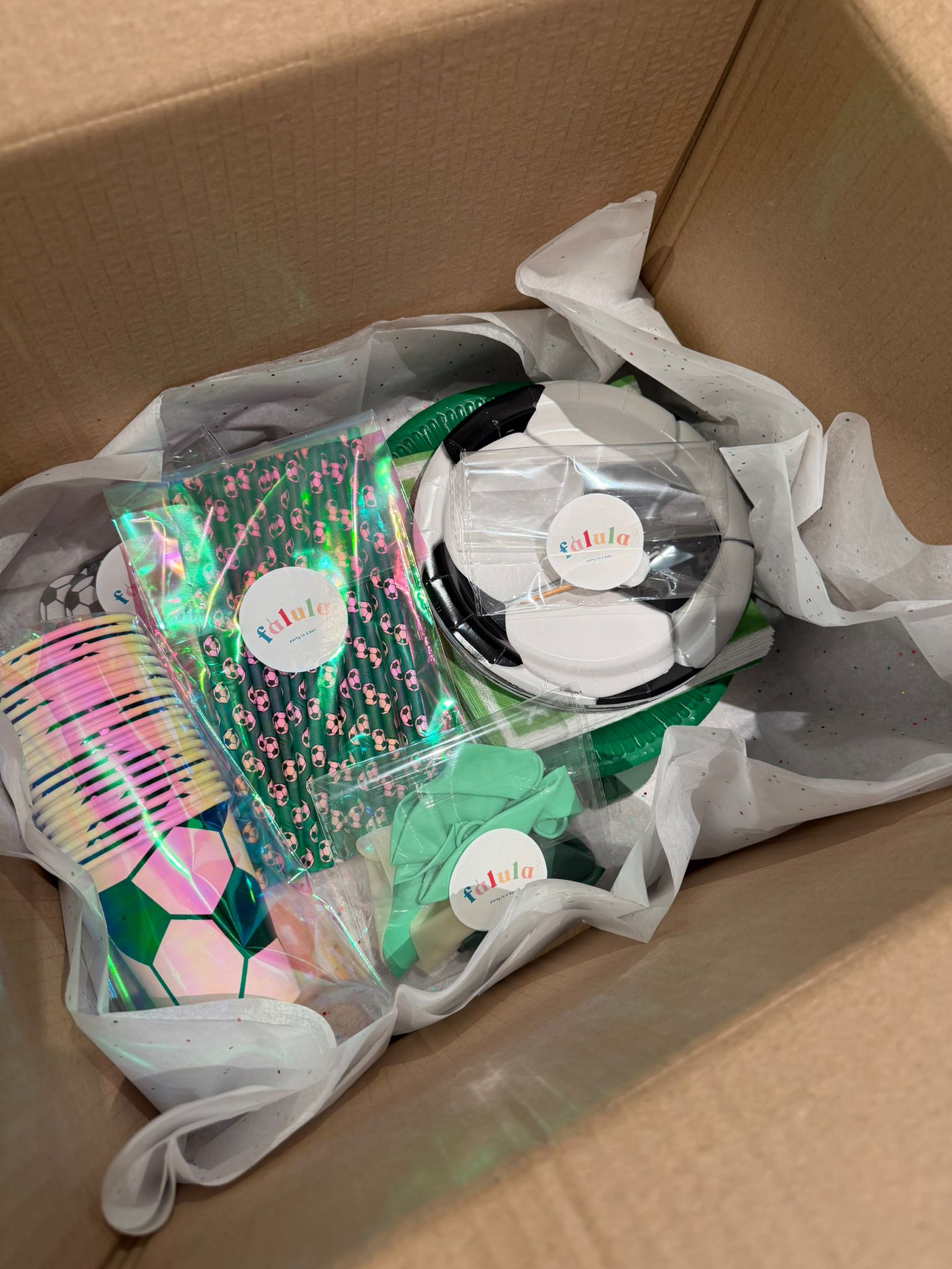 Caja de cartón con el pack completo de decoración para fiesta de fútbol, incluyendo platos de balones, vasos desechables, servilletas de campo de juego y velas para el cumpleaños.