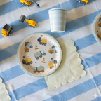 Primer plano de un plato de papel con temática de camiones de construcción, servilleta amarilla pastel con borde ondeado y vasos de cuadros azules sobre un mantel de rayas azul y blanco.