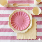 Primer plano de un servicio de mesa infantil compuesto por un plato rosa pastel sobre un plato base crema con ondas, una servilleta troquelada, un vaso de papel y una cuchara de madera desechable sobre un mantel de rayas rosas.
