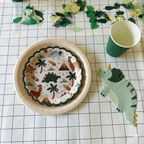 Primer plano de un plato de papel con ilustraciones de dinosaurios prehistóricos, una servilleta con forma de triceratops y un vaso verde sobre mantel geométrico.