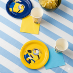 Primer plano de un plato amarillo y otro azul con temática de Minions, servilletas de papel de colores vibrantes y vasos a juego sobre un mantel de rayas azules y blancas.