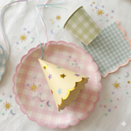 Primer plano de menaje para fiesta infantil de hadas con plato rosa de cuadros vichy, vaso verde pastel y gorrito de fiesta color crema con estrellas doradas y bordes festoneados sobre mantel blanco.