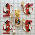Vista superior completa de la decoración de mesa para fiesta infantil con temática de coches vintage, mostrando cuatro juegos de platos con forma de coche, vasos de rayas, servilletas y dos cajas de palomitas y caramelos.