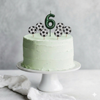 Tarta de cumpleaños infantil con cobertura de crema verde pastel, decorada con una vela metalizada con el número 6 y velas redondas con diseño de balones de fútbol tridimensionales.