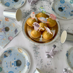 Detalle de mesa dulce con magdalenas decoradas con toppers de cohetes y astronautas sobre un plato plateado, rodeado de menaje de fiesta espacial.