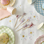 Vista aérea de una mesa de cumpleaños decorada con toppers para magdalenas en forma de hadas mágicas, rodeados de platos y vasos de colores pastel con estampado vichy y confeti de estrellas