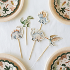 overhead view of five distinct wooden food picks featuring different cartoon dinosaurs (T-Rex, Triceratops, Brontosaurus, Stegosaurus, Pterodactyl) arranged on a white tablecloth with partial plates