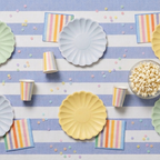 Vista aérea de una mesa decorada para fiesta infantil con platos ondulados en tonos pastel, vasos y servilletas a rayas de colores, acompañados de cuencos de cristal con palomitas y macarons sobre un mantel de rayas azules y blancas con confeti.