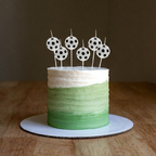 Tarta de cumpleaños decorada con seis velas con forma de balón de fútbol Falula, sobre un glaseado degradado verde y blanco y una mesa de madera.